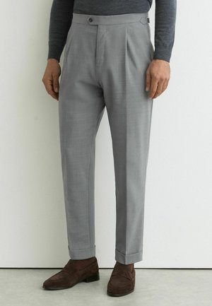 Muž oblečený v svetlosivých na mieru šitých nohaviciach a hnedých semišových mokasínach stojaci pri bielej stene na sivom podlaží.