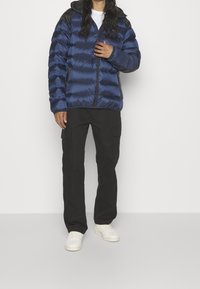 Marinblå pufferjacka med svarta detaljer, med dragkedja fram och huva, tillsammans med svarta cargobyxor och ljusa sneakers.