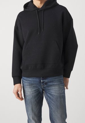 Homme portant un sweat à capuche noir tacheté avec poche avant et un jean bleu délavé, debout devant un fond uni.