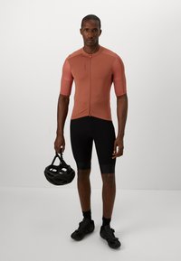 Maglia da ciclismo a maniche corte di colore ruggine con zip frontale, abbinata a pantaloni corti imbottiti neri. La persona tiene un casco nero.