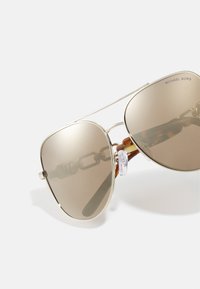 Occhiali da sole Michael Kors con lenti marroni sfumate, montatura in metallo argento e dettagli in tartaruga sulle tempie. Dettaglio a catena sul ponte del naso.