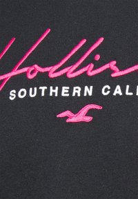 Svart tyg med rosa broderad text "Hollister" och "SOUTHERN CALI" samt en accentfågel-logotyp, som visar en stark färgkontrast och textur.
