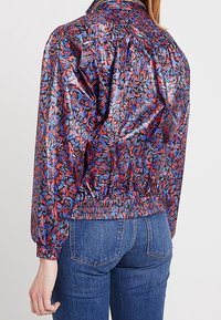 Veste coupe-vent colorée et à motifs, fabriquée dans un matériau léger et brillant, avec des poignets et une ceinture élastiqués. La vue arrière montre des coutures froncées.