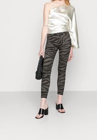 Jednostranný metalický top v béžové barvě, doplněný o úzké olivově zelené kalhoty s černým zebrovaným vzorem a černými sandály na blokovém podpatku.