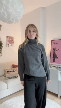 Jersey de cuello alto gris y pantalones de pierna ancha negros. El jersey tiene una textura suave; los pantalones están hechos de un material ligero. Se puede ver la decoración de la habitación.