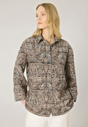 Femme aux cheveux blonds portant une chemise boutonnée à manches longues avec un motif floral beige et marron, debout les mains dans les poches d’un pantalon blanc.