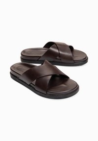 Paire de sandales en cuir brun foncé avec des larges brides croisées et des semelles noires rembourrées sur fond blanc.