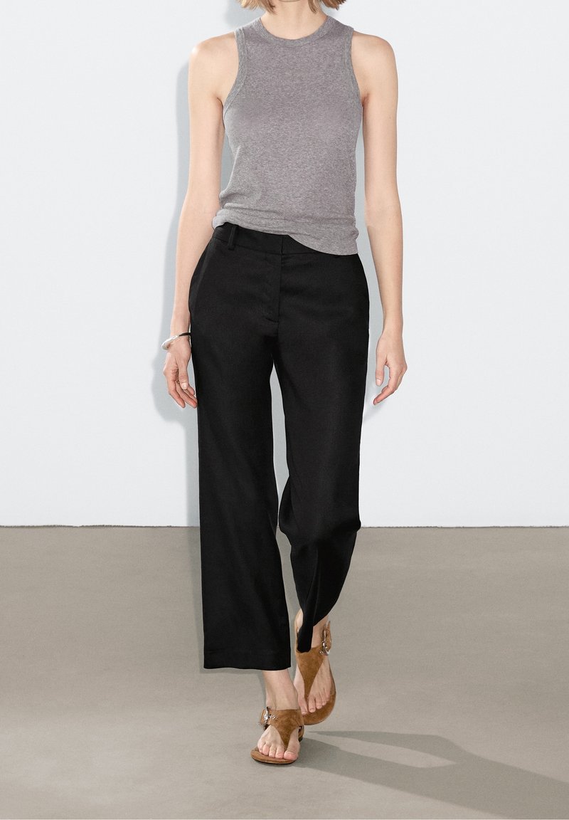 Femme portant un haut gris sans manches, un pantalon noir à jambes larges et des sandales marron ouvertes aux orteils, debout sur un sol gris contre un mur blanc.