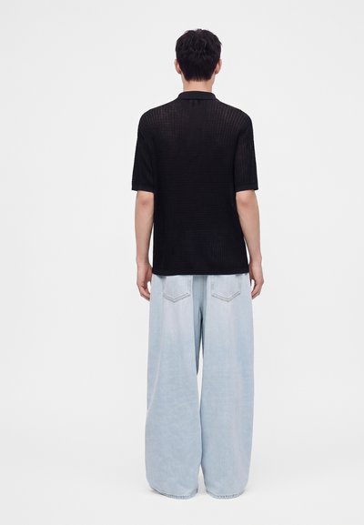 Personne portant un t-shirt noir à manches courtes et un jean large bleu clair, debout dos à la caméra sur un fond blanc uni.