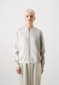 Helles graues Zip-Hoodie mit hohem Kragen, gerippten Bündchen und Seitentaschen, aus glattem Stoff gefertigt. Über lockeren beigen Hosen getragen.