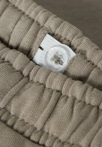 Beżowa dzianina o pikowanej fakturze i poziomych plisach. Posiada biały elastyczny pasek oraz przezroczysty guzik z kwiatowym wzorem.