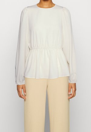 Vrouw die een witte blouse met lange mouwen en een geplooide taille draagt, gecombineerd met beige, hooggetailleerde, wijde broekspijpen, rechtopstaand tegen een effen achtergrond.