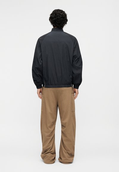 Isik lokkis tume juustega, kandes musta bomberjopet ja lahtisi pruune pükse, seisab seljaga vaataja poole ühtlase tausta ees.