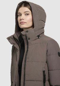 Smeđa podstavljena jakna s kapuljačom, koja ima prednji patentni zatvarač, dugmad na preklop i specifične šavove. Uključuje bočne džepove s patentnim zatvaračem.