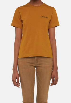Złota bawełniana koszulka z krótkim rękawem i okrągłym dekoltem, z haftem "TRUSSARDI" w czerni na lewej piersi. Noszona z beżowymi spodniami.