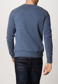 Sudadera azul hecha de un tejido suave, con cuello redondo, mangas largas y puños acanalados. Se lleva con jeans oscuros que muestran detalles en los bolsillos traseros.