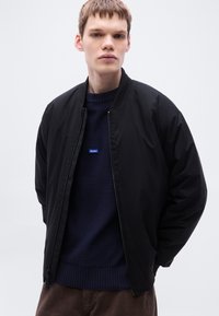 Μαύρο μπουφάν bomber με φερμουάρ μπροστά, ριμπ κολάρο και μανσέτες, φορεμένο πάνω από μπλε πουλόβερ με μικρό μπλε λογότυπο "HUGO".