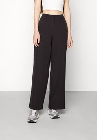 Person som bär svarta vida byxor med hög midja, vit kort topp och silverfärgade sneakers, stående mot en enfärgad vit bakgrund.