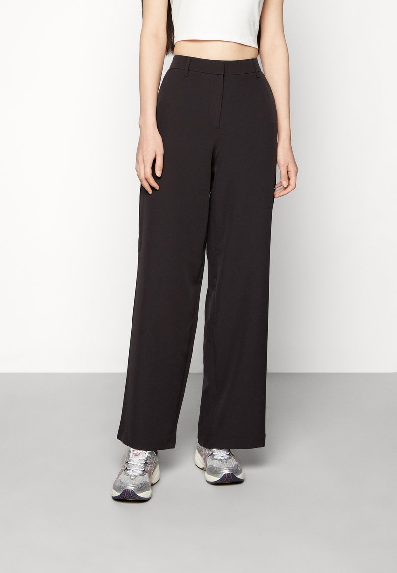 Person som bär svarta vida byxor med hög midja, vit kort topp och silverfärgade sneakers, stående mot en enfärgad vit bakgrund.