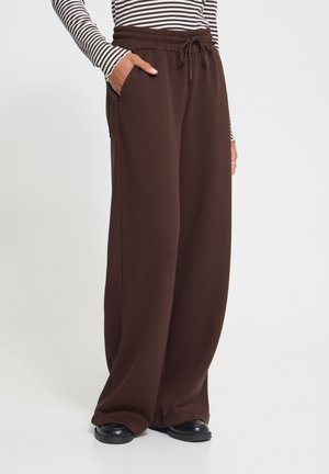 Persona che indossa pantaloni larghi marroni con coulisse, mani nelle tasche, scarpe nere e una maglia a maniche lunghe a righe bianche e nere.