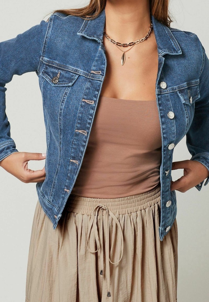 Chaqueta vaquera azul medio con un corte entallado, que presenta botones a presión plateados, dos bolsillos en el pecho y costuras en contraste. Mangas largas.