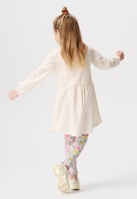 Robe blanche en broderie anglaise avec des manches longues et une taille cintrée, associée à des leggings à motifs floraux et des baskets claires.