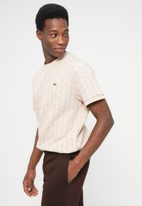 Joven con camisa de manga corta beige con estampado y logo, pantalones oscuros, mirando de lado con una mano en el bolsillo, contra un fondo liso.