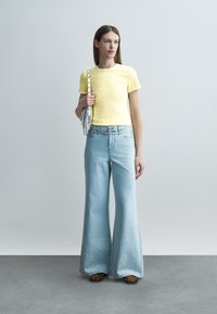 Mujer de pie con camiseta de manga corta de color amarillo claro, vaqueros anchos de pierna en azul claro, zapatos con estampado de leopardo, sosteniendo un bolso blanco con flecos al hombro.