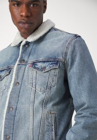 Veste en denim bleu clair avec un col en shearling crème. Comprend deux poches poitrine, des boutons argentés et des coutures contrastantes le long des coutures.