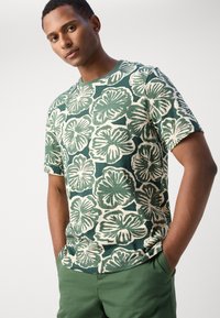 Green floral patterned t-shirt with short sleeves, round neckline, and beige accents. Worn with green pants, showcasing a relaxed fit.