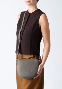 Šedá kožená kabelka přes rameno s kulatým tvarem, embosované logo "JOOP! JEANS", odnímatelný popruh a strukturovaný povrch, vystavená na minimalistickém pozadí.