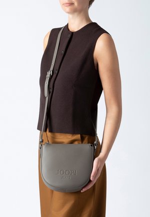 Šedá kožená kabelka přes rameno s kulatým tvarem, embosované logo "JOOP! JEANS", odnímatelný popruh a strukturovaný povrch, vystavená na minimalistickém pozadí.