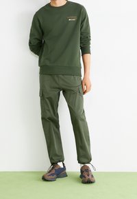 Grön huvtröja och cargo byxor, båda i matchande färg. Huvtröjan har ribbade mudd armar och en logotyp i ljusare grön. Bruna och marinblå sneakers.