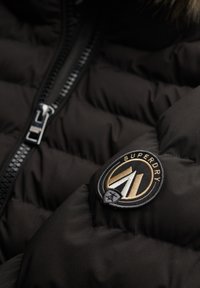 Chaqueta acolchada negra con un primer plano de una cremallera metálica y un parche redondo con el logo de Superdry en dorado, blanco y negro en la manga.