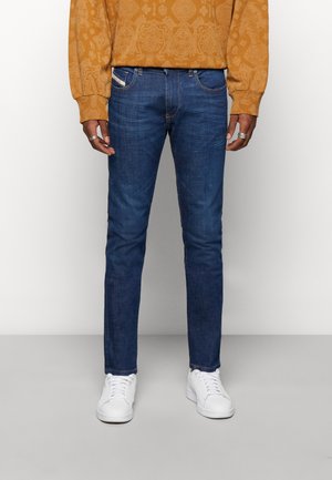 Jeans in denim blu scuro con vestibilità slim, accentuati da cuciture gialle, tasche frontali e una leggera sfumatura. Indossati con scarpe da ginnastica bianche.