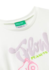 Bílý krátký rukáv tričko s velkým pastelově fialovým nápisem "Glow" a růžovou kreslenou postavičkou, se zeleným logem "PEANUTS" níže.