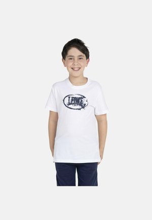 Lächelnder Junge stehend, trägt ein weißes Leone 1947 T-Shirt und dunkle Shorts vor einem einfarbigen hellen Hintergrund.