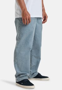 Jean en denim bleu clair avec une coupe décontractée, présentant une jambe droite et une texture subtile. Porté avec un t-shirt blanc et des baskets sombres.