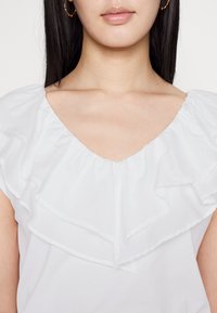 Blouse blanche avec un décolleté en V et un détail de volants superposés aux épaules. Texture lisse avec un léger éclat, design court.