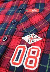 Chemise en flanelle à carreaux rouge et marine avec des boutons noirs, broderie "08" et un patch Union Jack sur le devant. Texture douce, design décontracté.