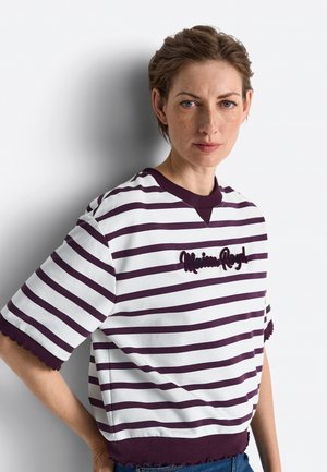 Vrouw met kort haar die een wit en donkerpaars gestreept shirt met geborduurde tekst draagt, staand tegen een lichte achtergrond.