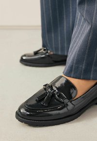 Schwarze Lackleder-Loafer mit Quasten und einer Kettenakzentuierung; strukturierte Oberfläche; runde Zehenform; kombiniert mit gestreiften Hosen.