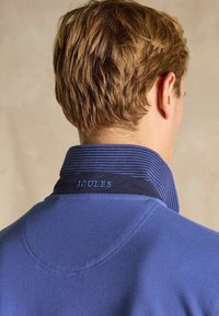 Zils džemperis ar ribotu apkakli. Satur vienkāršu dizainu, teksturētu audumu un uzrakstu "JOULES" izšūtu gaiši zilā krāsā uz apkakles.