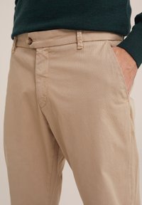 Beige katoenen broeken met zichtbare stiksels, voorzakken en een knoopsluiting in de taille. Plat voorpand ontwerp, gladde textuur.