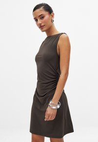 Robe marron sans manches en tissu lisse. Présente un design drapé avec des détails froncés sur le côté. Accessoirisée avec un bracelet transparent.