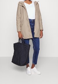 Beige waterdichte jas met capuchon, wit t-shirt, hoge blauwe jeans en zwarte gewatteerde schoudertas. Witte sneakers maken de look compleet.