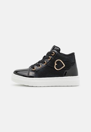 Baskets montantes noires avec semelle blanche, boucle dorée en forme de cœur et lacets noirs sur fond clair uni.