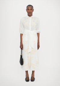 White linen dress with light beige floral embroidery, fitted waist with a tie, short puff sleeves, paired with a black textured bag.