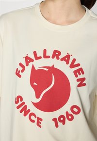 Krém színű pamut póló, amelyen egy nagy piros köralakú logó található, amelyen egy róka és a "FJÄLLRÄVEN SINCE 1960" felirat olvasható vastag betűtípussal. Kerek nyakú kivitel.