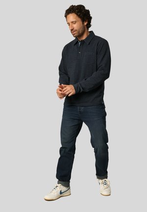 Pull en marine avec un col et une poche sur la poitrine, associé à un jean denim foncé et des baskets blanches avec des accents bleus.